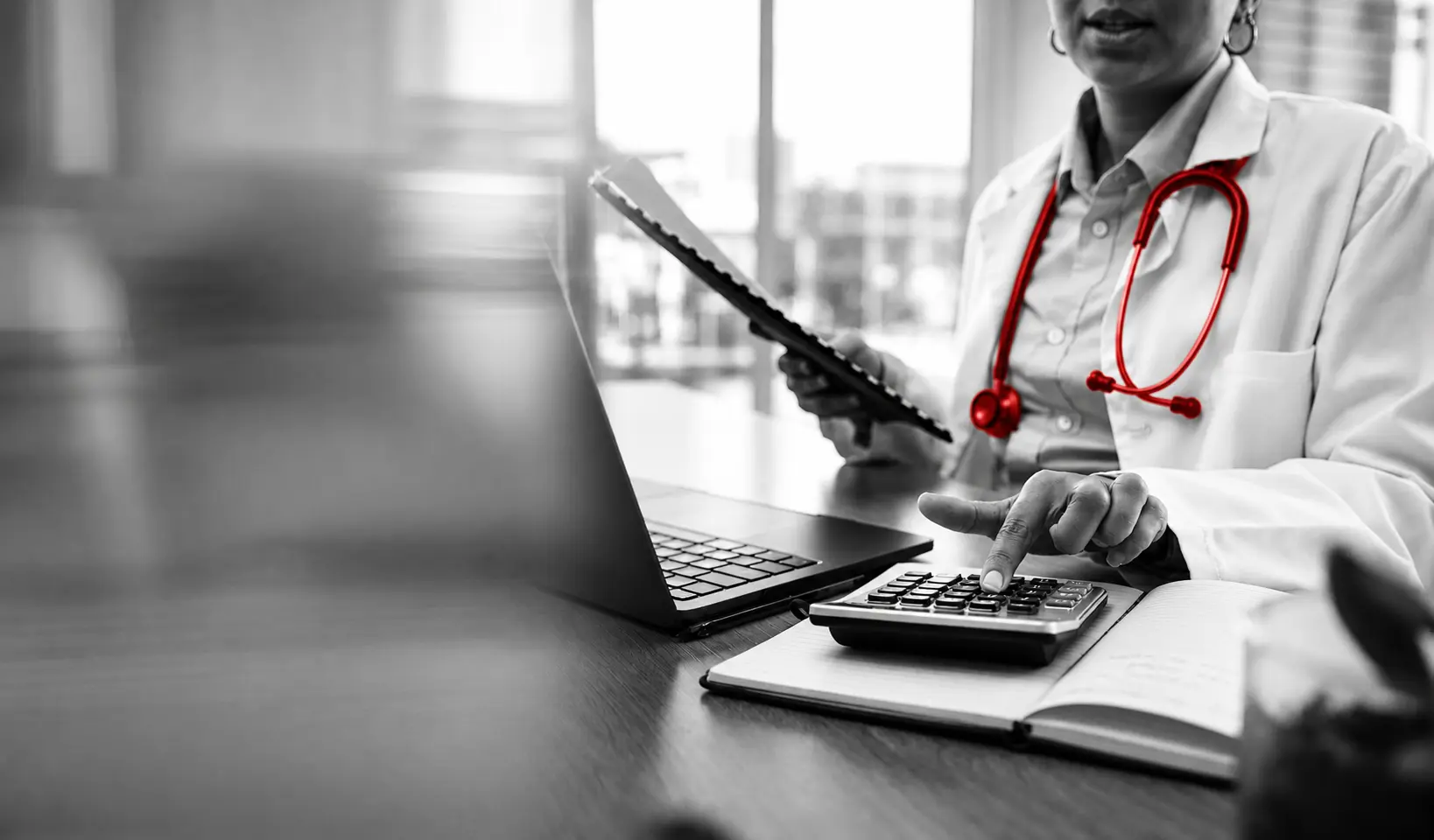 A imagem mostra em destaque as mãos de uma médica, sentada à mesa, em seu consultório. Com uma das mãos, ela segura uma planilha e, com a outra, faz cálculos em uma calculadora.
