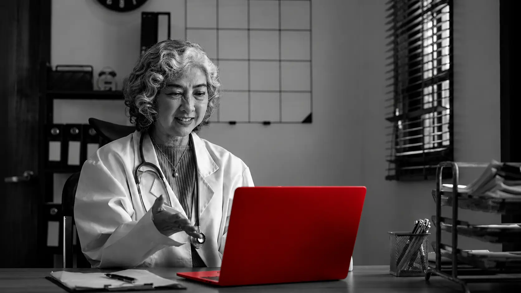 A imagem mostra uma médica de meia-idade, usando jaleco e estetoscópio em volta do pescoço, sentada à mesa em seu consultório, fazendo uma consulta remota em seu laptop.