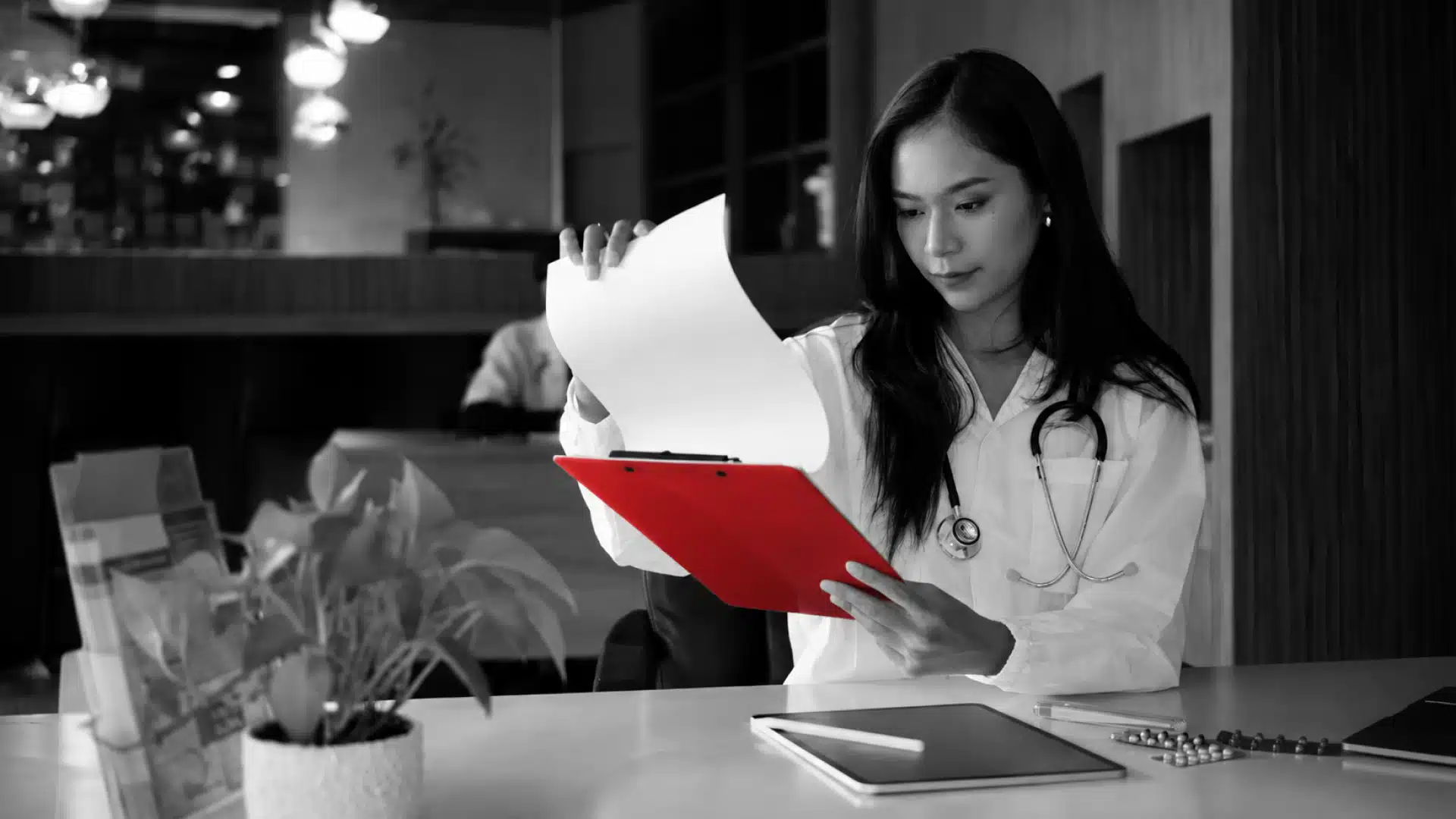 A imagem mostra uma médica, sentada à mesa, em seu consultório, analisando papéis.