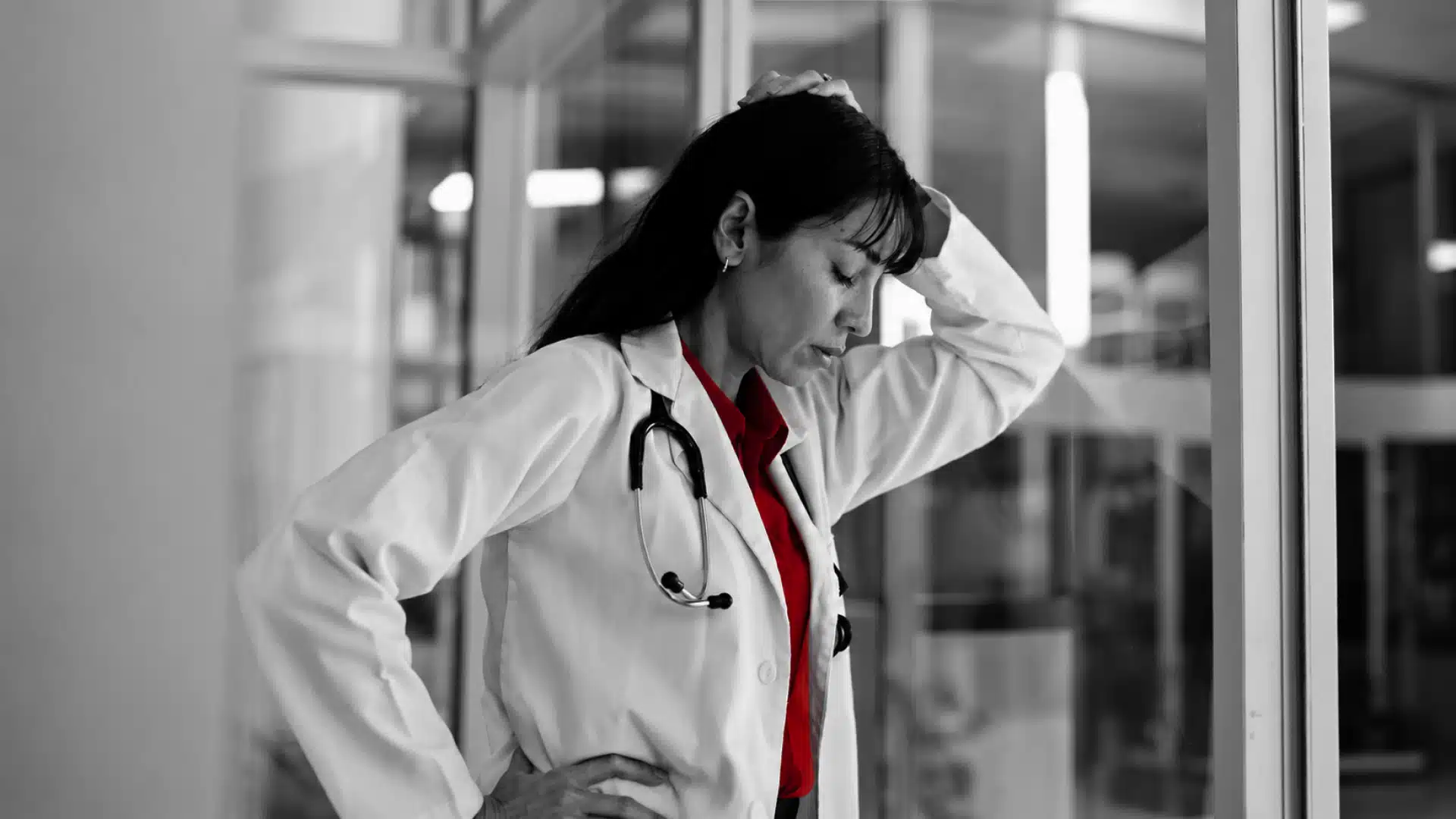 A imagem mostra uma médica, usando jaleco, estetoscópio, em ambiente hospitalar com uma expressão preocupada.